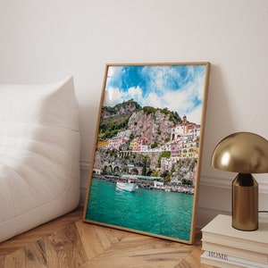 May include: A framed photograph of a colorful town built into a cliffside overlooking a turquoise body of water. The town is surrounded by lush green vegetation and a bright blue sky with fluffy white clouds.