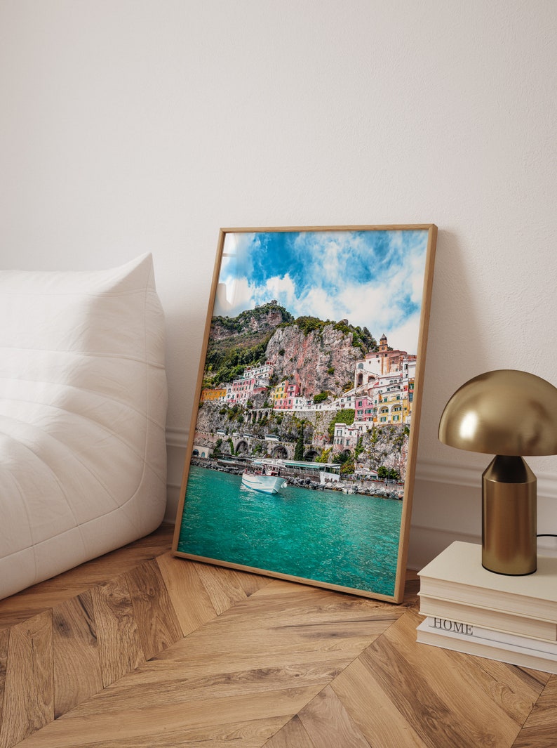 May include: A framed photograph of a colorful town built into a cliffside overlooking a turquoise body of water. The town is surrounded by lush green vegetation and a bright blue sky with fluffy white clouds.