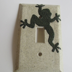 Puede incluir: Una tapa de interruptor de luz de cerámica beige claro con una silueta de rana verde oscura.