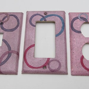 Puede incluir: Juego de tres cubiertas decorativas para interruptores de luz. Las cubiertas son de color rosa moteado con círculos superpuestos en tonos morado, azul y rojo. Una cubierta tiene una abertura para un interruptor, otra para una toma doble y la tercera para una toma simple.