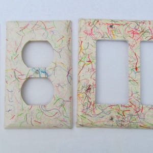 May include: A set of decorative light switch covers and outlet covers. The covers have a cream-colored background with colorful, thin, random lines of red, green, yellow, and blue. The set includes a single switch cover, a double outlet cover, and a triple switch cover.