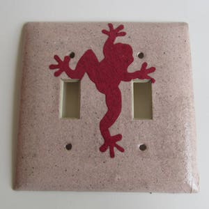 Puede incluir: Una tapa de interruptor de luz doble de color marrón claro con un diseño de silueta de rana roja.