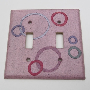 Puede incluir: Placa decorativa para interruptor de luz con fondo rosa y círculos coloridos superpuestos. Los círculos son en tonos morado, rojo y gris. La placa tiene dos aberturas para interruptores y orificios para tornillos.