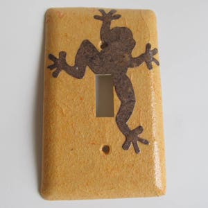 Puede incluir: Una tapa de interruptor de luz con un fondo amarillo texturizado y una silueta de rana marrón.