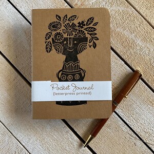 Peut inclure: Un journal de poche marron avec un motif floral noir et les mots "Pocket Journal" imprimés sur une bande blanche. Un stylo marron avec des accents dorés est posé à côté du journal. Le journal est sur une surface en bois.