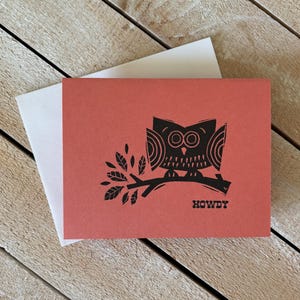 Peut inclure: Une carte de vœux corail avec une illustration de hibou noir perché sur une branche, accompagnée d'une enveloppe blanche. Le mot "HOWDY" est imprimé en dessous du hibou. La carte est posée sur un fond en bois.