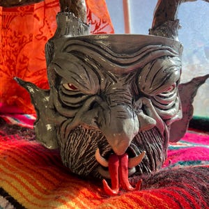 May include: A gray, devil-faced ceramic planter with red eyes, horns, and a protruding red tongue. The planter has a textured surface and is set against a colorful, patterned textile.