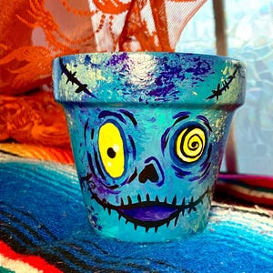 May include: A hand-painted teal ceramic flower pot with a zombie face design. The zombie face has yellow eyes, a black nose, and a purple mouth. The pot is decorated with black stitches and purple accents.