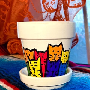 May include: White ceramic flower pot with a matching saucer. The pot is decorated with a colorful, cartoon-style illustration of several cats in various colors, including yellow, orange, red, and blue. The pot is sitting on a colorful blanket.