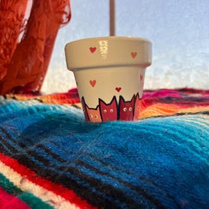 May include: A small, white ceramic flower pot with a hand-painted design. The pot features pink hearts around the rim and a row of stylized cat silhouettes in shades of pink and red. The pot is set on a colorful, striped blanket.