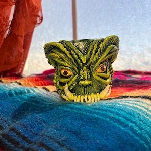 May include: A green and yellow monster-themed ceramic planter with red eyes and white teeth. The planter is sitting on a colorful striped blanket with blue and red hues. The planter has a detailed, textured design.