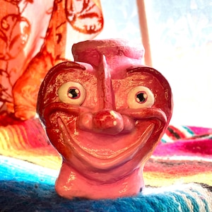 May include: A pink ceramic face sculpture with a smiling expression, large eyes, and a prominent nose. The sculpture is set against a colorful textile background and a sheer orange curtain.