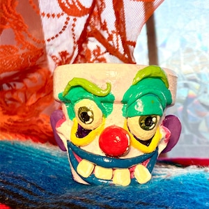 May include: A hand-painted ceramic clown face planter. The planter features a white base with green eyebrows and hair, large yellow eyes, a red nose, and a blue mouth with white teeth. The background includes orange lace and a blue and red striped textile.