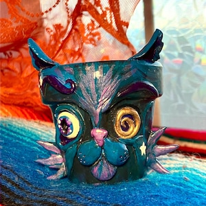 May include: A hand-painted teal ceramic planter shaped like a cat's face. The planter features large, expressive eyes, a pink nose, and a whimsical design with purple and gold accents. The cat has pointed ears and star details.