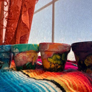 May include: Four small, hand-painted terracotta flower pots. Each pot features a unique landscape design with trees, clouds, and a house, in shades of blue, green, orange, and purple. The pots are displayed on a colorful, patterned blanket.