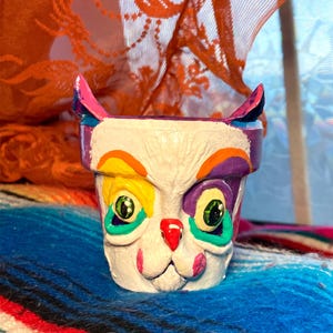 May include: A hand-painted, white ceramic planter shaped like a cat's face. The cat has colorful accents, including yellow, purple, and green. The planter is sitting on a colorful, striped blanket. The background includes orange lace fabric.