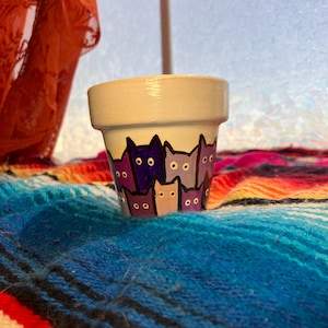 May include: A small, off-white ceramic flower pot with a painted cat design. The cats are in shades of purple and gray, with black outlines. The pot sits on a colorful, striped blanket.