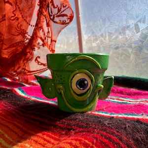 May include: A small, green ceramic planter with a whimsical monster face design. The planter features a large, blue and white eye, a smiling mouth, and two small ears. The planter is sitting on a colorful, striped blanket.