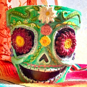 May include: Hand-painted ceramic flower pot in the shape of a sugar skull. The pot is painted in shades of green, with floral accents and large, vibrant red and yellow flower eyes. The mouth is open, revealing a dark interior.