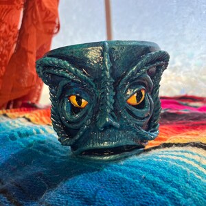 May include: A teal-colored, sculpted ceramic pot with a creature's face. The face has large, yellow eyes, a prominent nose, and a detailed, textured surface. The pot is sitting on a colorful, patterned blanket.