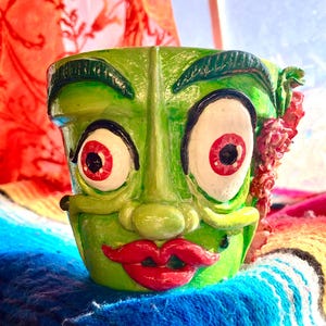 May include: A hand-painted, green ceramic planter with a whimsical face design. Features large, red-eyed eyeballs, a red mouth, and decorative details. The planter is set against a colorful, textured background.