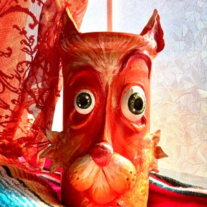 May include: A whimsical, orange ceramic cat-faced vase with large green eyes and pointed ears. The vase has a cylindrical shape and is decorated with a stylized cat face. The background includes a red curtain and a colorful textile.
