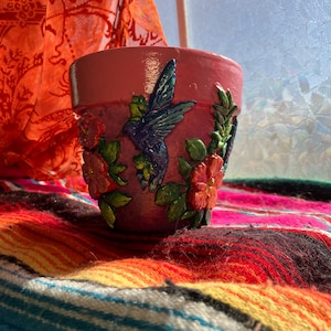 May include: A hand-painted terracotta flower pot in a dusty rose color. The pot features a detailed relief of a hummingbird in flight, surrounded by red flowers and green leaves. The pot is sitting on a colorful woven blanket.