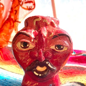 May include: A red ceramic vase with a face design. The vase has a detailed face with dark eyes, a black mustache, and a smiling mouth. The vase is painted in shades of red and has a glossy finish. The background includes colorful textiles.