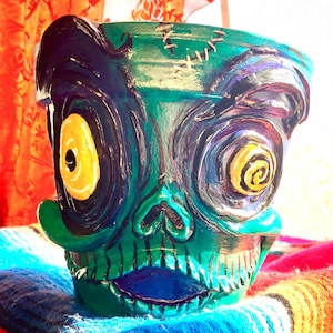May include: A hand-painted ceramic planter in the shape of a zombie skull. The planter features green, blue, and yellow paint with stitched details. The eyes are yellow spirals, and the mouth is blue.