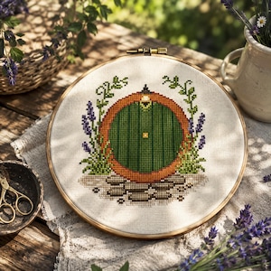 May include: Cross-stitch embroidery hoop featuring a green door with an orange frame, surrounded by lavender and greenery. The design is set against a cream-colored fabric, framed by a wooden hoop. The scene evokes a cozy, whimsical aesthetic.