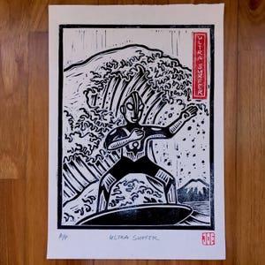 Puede incluir: Grabado en madera en blanco y negro de un superhéroe surfeando una gran ola. La impresión se titula "Ultra Surfer" y está firmada por "Joe".