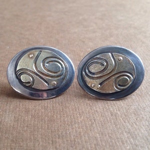 Op de afbeelding: Een paar zilveren oorbellen met een gouden swirl-ontwerp. De oorbellen zijn ovaalvormig en hebben een gladde, gepolijste afwerking.