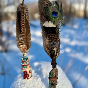 May include: Two decorative feather wands. One wand features a totem-like handle with a colorful design. The other wand has a peacock feather with a blue and green eye. Both wands are adorned with beads and other decorative elements.