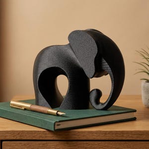 May include: A matte black elephant figurine with a textured surface and a curved trunk, positioned on a dark green book next to a gold and wood fountain pen. The elephant has a solid, stylized form with a large ear and a rounded body shape.