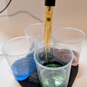 May include: A black plastic holder with four clear plastic cups filled with blue, green, and pink liquids. A gold probe is inserted into the holder.