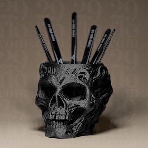 Puede incluir: Un portalápices negro con forma de calavera con una superficie texturizada detallada. Varios bolígrafos negros están colocados dentro del soporte. El portalápices es un artículo decorativo, adecuado para un escritorio o espacio de trabajo.