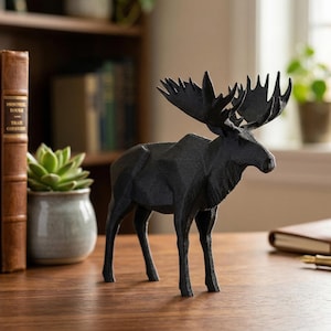 Puede incluir: Una figura de alce negro con grandes astas sobre una superficie de madera. La escultura es detallada y realista, mostrando la forma del animal. El fondo incluye una estantería, una planta en maceta y un cuaderno.