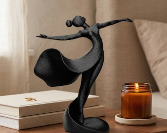 May include: A black sculpture of a dancing figure with arms outstretched and a flowing skirt. The figure has a textured surface and is captured in a dynamic pose with one leg extended, creating a sense of movement and grace in this decorative piece.