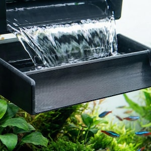 May include: A black aquarium waterfall filter with water cascading into a rectangular basin. The filter is mounted above lush green aquatic plants and small colorful fish, creating a naturalistic underwater scene.