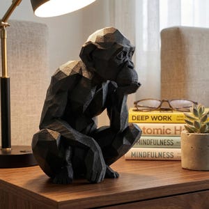 Può includere: Una scultura di scimpanzé geometrico nero in posizione seduta, con il mento appoggiato sulla mano. La scultura è su una superficie di legno, con una lampada, libri e una piccola pianta in vaso sullo sfondo.