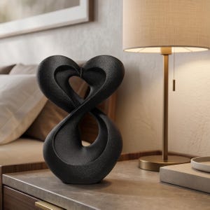 Puede incluir: Una escultura negra con superficie texturizada que presenta un diseño abstracto de bucle continuo que forma dos figuras interconectadas similares a corazones. El objeto tiene una silueta suave y curva y se mantiene en posición vertical.