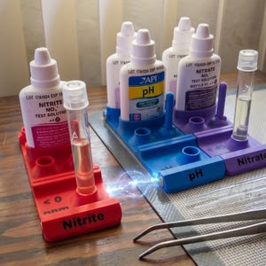 May include: A collection of aquarium water test kits. The image shows bottles of test solutions, test tubes, and holders in red, blue, and purple. The bottles are labeled with text including "Nitrite", "pH", and "Nitrate".