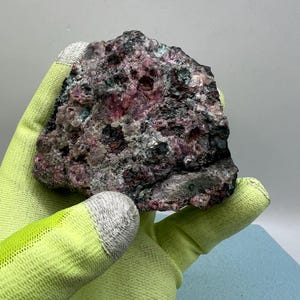 May include: A rough, multi-colored mineral specimen. The rock features a mix of pink, gray, green, and black hues. The mineral is held by a person wearing a yellow-green glove.