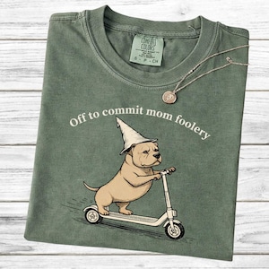 Może przedstawiać: Szałwiowa zielona koszulka z napisem "Off to commit mom foolery" i rysunkowym psem w kapeluszu czarodzieja jadącym na skuterze. Widoczny jest również złoty naszyjnik z wisiorkiem.