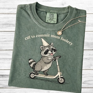 Może przedstawiać: Zielona koszulka Comfort Colors z napisem "Off to commit mom foolery" i grafiką szopa jadącego na skuterze. Widoczny jest również złoty naszyjnik z wisiorkiem.