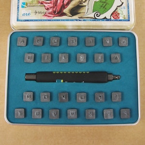 Puede incluir: Una caja de metal con un interior azul que contiene un juego de sellos de letras y números. Los sellos están dispuestos en filas y son de metal. Se incluye una herramienta de metal negro en la caja.
