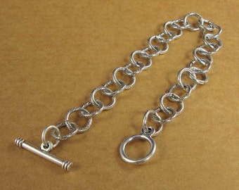 May include: A silver chain bracelet with a toggle clasp and a ring closure. The chain is made of twisted links.