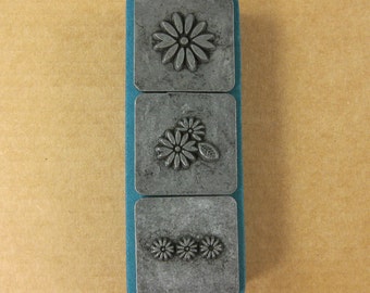 May include: A metal stamp set with three rectangular stamps, each featuring a different floral design. The stamps are mounted on a teal blue block. The background is a light brown color.