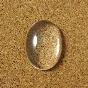 May include: A clear glass oval-shaped magnifying glass on a cork background.