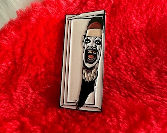 May include: A white enamel pin with a black outline depicting a man with a red Santa hat peeking out of a door frame. The man is smiling with his mouth open and his eyes wide. The pin is on a red fuzzy surface.